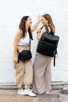 Two women stand side by side with hands in pockets, each wearing a black travel backpack.