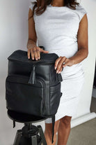 A woman in a white dress holds a suitcase and a black leather baby backpack