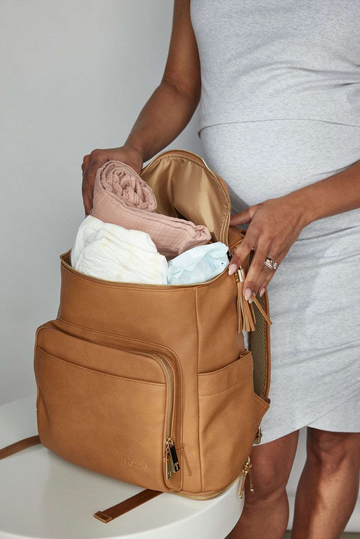 A pregnant woman holds an unzipped tan colour leather baby backpack