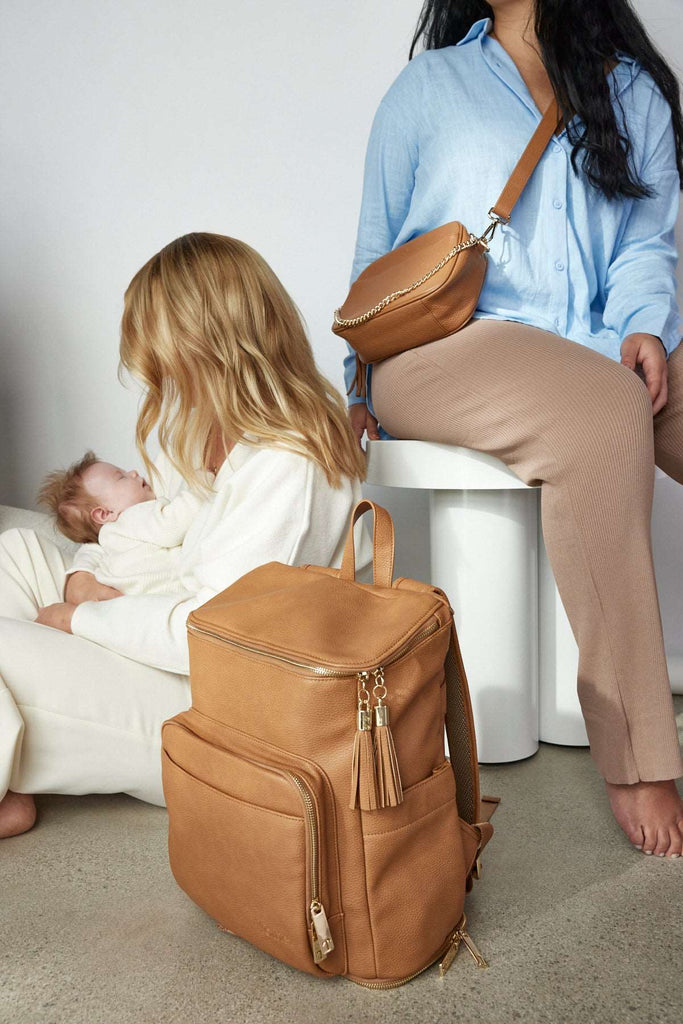 Two women sit on the floor with a baby and a tan colour leather baby backpack beside them