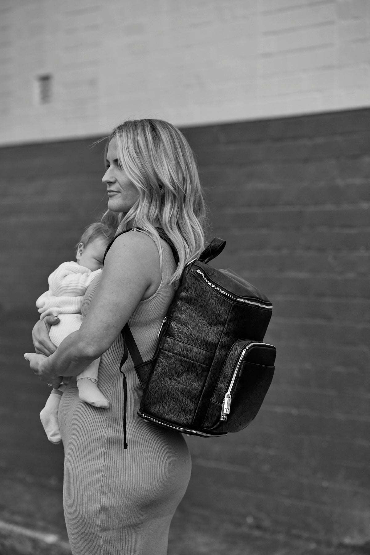 A black and white photo of a woman holding a baby, carrying a black leather baby backpack