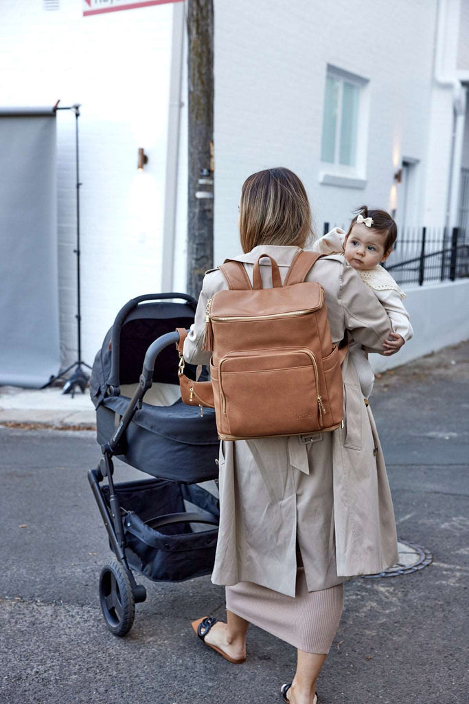 A woman walks down the street with a baby and pushing a stroller while carrying a tan colour leather baby backpack