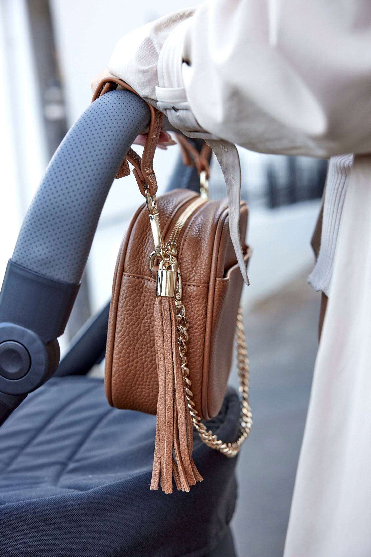 A woman pushing a stroller, with a tan colour bag with pram clips in gold hanging on the stroller