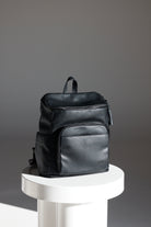 A black baby bag backpack rests on a white pedestal