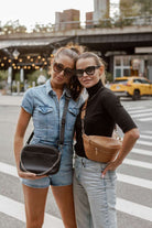 Two women stand on a bustling city street, one carrying a black Bestie Crossbody Bag and the other sporting a tan Bum Bag, both from Tottie.