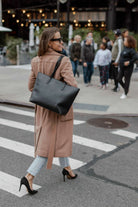 A woman in a coat and heels crosses the street while carrying a black tote bag